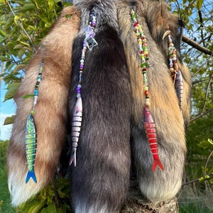 Peut inclure: Plusieurs queues de renard aux tons bruns, gris et fauves, ornées de brins perlés et de breloques en forme de poissons en bois peints. Les queues sont suspendues à un arbre, avec le mot "liberted" écrit sur une petite pancarte.