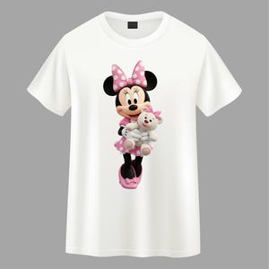 May include: A white short-sleeved t-shirt featuring a graphic print of Minnie Mouse wearing a pink polka-dot dress and matching bow, holding a small white teddy bear with a pink bow on its head. The character has black ears and pink shoes.