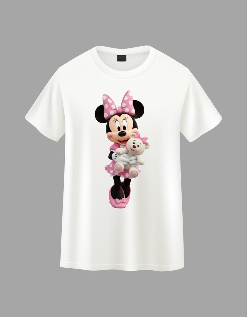 May include: A white short-sleeved t-shirt featuring a graphic print of Minnie Mouse wearing a pink polka-dot dress and matching bow, holding a small white teddy bear with a pink bow on its head. The character has black ears and pink shoes.