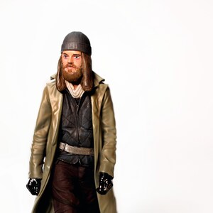 May include: A figurine of a man wearing a gray beanie, a long tan coat, and black gloves. The figure has a beard and is wearing a black quilted vest over a black shirt. The figure is standing against a white background.