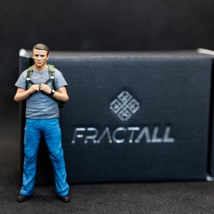 Op de afbeelding: Een miniatuurfiguur van een persoon in een grijs t-shirt, blauwe jeans en een rugzak staat voor een zwarte doos. De doos heeft het woord "FRACTALL" in zilveren letters en een geometrisch ontwerp.