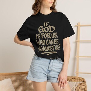 Könnte beinhalten: Ein schwarzes kurzärmeliges T-Shirt mit dem beigen Schriftzug "IF GOD IS FOR US, WHO CAN BE AGAINST US?" auf der Vorderseite, wobei der kleinere Text "ROMANS 8:31" direkt unter dem Hauptzitat in einer passenden beigen Farbe gedruckt ist.