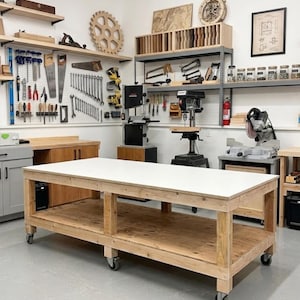 May include: A large rectangular wooden workbench with a smooth white top, supported by a sturdy frame with a lower shelf and six swivel caster wheels, positioned in a workshop filled with various power tools and hand tools on wall pegboards.