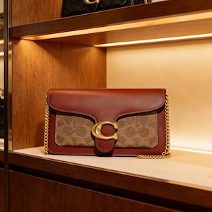 Op de afbeelding: Een bruine leren schoudertas met een gouden kettingriem en een gouden sluiting. De tas heeft een bruin en tan patroon op het voorpaneel en wordt tentoongesteld op een houten plank. Een andere tas is zichtbaar op de plank erboven.