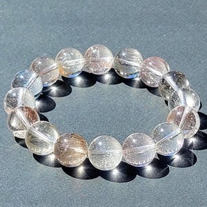 May include: A clear quartz bead bracelet with internal inclusions. The round beads are strung together, creating a circular shape. The bracelet is resting on a dark gray surface, with shadows cast by the sunlight.