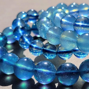 Op de afbeelding: Een armband bestaande uit doorschijnende, bolvormige blauwe kralen met een iriserende afwerking. De kralen hebben een reflecterende kwaliteit en vertonen subtiele verschuivingen in licht en kleur over hun gladde, afgeronde oppervlakken.
