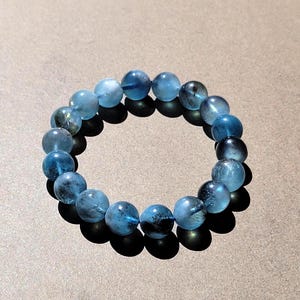 Könnte beinhalten: Ein kreisförmiges Armband aus durchscheinenden blauen Perlen. Jede Perle weist dunkle Einschlüsse und verschiedene Schattierungen von hellem bis tiefem Blau auf, was ein marmoriertes Aussehen erzeugt. Die Perlen sind zu einer Schlaufe aufgereiht.