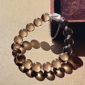 Könnte beinhalten: Ein Armband bestehend aus runden, durchscheinenden, rauchbraunen Quarzperlen, die aneinandergereiht sind. Das Armband verfügt über einen silberfarbenen Metallverschluss mit einer kleinen, feinen Sicherheitskette, um die Enden des Strangs zu sichern.