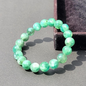 May include: A circular bracelet composed of smooth, round, translucent green beads with internal white marbling and inclusions. The beads are strung together in a continuous loop, displaying a polished finish that reflects light across the surface.