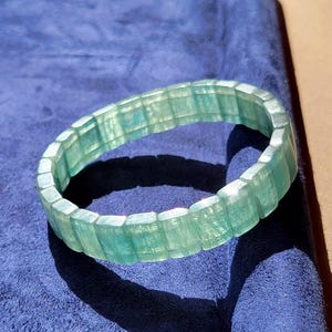 Könnte beinhalten: Ein durchscheinendes hellgrünes Armband, das aus rechteckigen, polierten Segmenten besteht, die in einem durchgehenden kreisförmigen Band angeordnet sind. Das Material weist eine feine, faserige Innenstruktur mit Nuancen von Minzgrün auf.