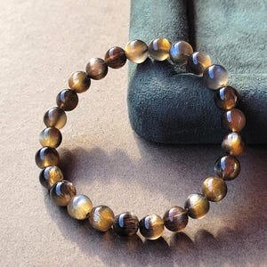 Può includere: Un bracciale circolare composto da perline sferiche levigate in tonalità marrone, oro e grigio traslucido. Le perline presentano inclusioni fibrose interne che creano un effetto scintillante e chatoyant quando la luce colpisce le superfici lisce.