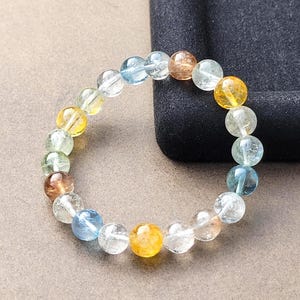 Könnte beinhalten: Ein kreisförmiges Armband aus glatten, runden, durchscheinenden Edelsteinperlen in verschiedenen Farben, darunter klar, hellblau, blassgrün und goldgelb. Die Perlen sind auf ein elastisches Band aufgezogen und bilden eine geschlossene Schleife.