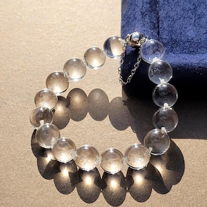 May include: A bracelet composed of clear, spherical beads arranged in a circular pattern. The beads are transparent and reflect light, creating distinct shadows. A metallic clasp with a small attached safety chain secures the ends of the bracelet.