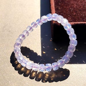 Puede incluir: Una pulsera elástica compuesta por cuentas translúcidas, facetadas y de color morado claro con un brillo iridiscente. Las cuentas están dispuestas en un patrón circular, reflejando la luz para crear sutiles destellos en sus superficies pulidas.