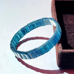 May include: A bracelet composed of rectangular, translucent blue beads arranged in a continuous circular band. The beads exhibit varying shades of deep and light blue, creating a layered, crystalline appearance throughout the entire piece.