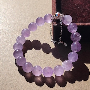 Op de afbeelding: Een armband bestaande uit gladde, ronde, lichtpaarse amethistkralen. De armband is voorzien van een zilverkleurige metalen sluiting en een kort, delicaat zilverkleurig kettingverlengstuk voor een verstelbare pasvorm.