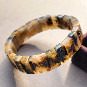 以下が含まれることがあります： 磨かれた琥珀色の長方形のビーズで構成された伸縮性のあるブレスレット。各ビーズには、シダのような独特の暗いインクルージョンが含まれており、半透明の黄金色の素材全体に枝分かれした有機的なパターンを作り出しています。