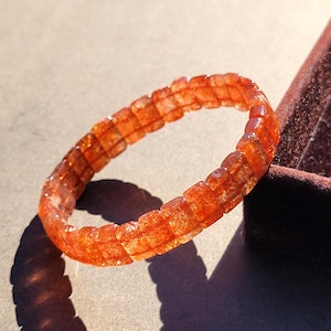 Può includere: Un braccialetto composto da piccole perline rettangolari in pietra del sole arancione traslucida. Le perline presentano una consistenza interna scintillante e brillante con varie sfumature di arancione intenso e marrone rossastro lungo tutto il cerchio.