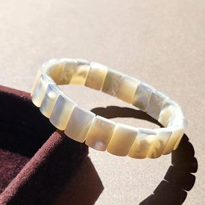 Op de afbeelding: Een cirkelvormige armband bestaande uit individuele, rechthoekige, gebroken witte, iriserende schelpsegmenten die met elkaar zijn verbonden. De segmenten hebben een gladde, gepolijste afwerking met subtiele kleurvariaties.