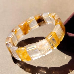 Op de afbeelding: Een elastische armband bestaande uit afwisselende rechthoekige kralen. De kralen bevatten een mix van doorschijnende heldere delen en levendige goudgele insluitingen, wat een herhalend patroon van lichte en donkere tinten in de band creëert.