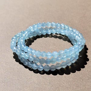 May include: A triple-strand bracelet composed of small, round, translucent light blue beads. The beads have a smooth, polished finish and are strung together in a circular arrangement, creating a layered appearance with a consistent pale blue hue.