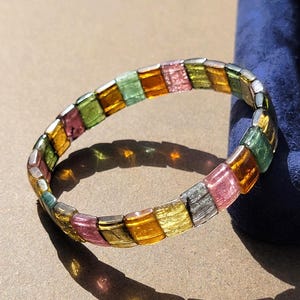 Op de afbeelding: Een cirkelvormige armband bestaande uit kleine, rechthoekige, doorschijnende glaskralen in afwisselende tinten amber, roze, groen en zilver. De kralen zijn gerangschikt in een herhalend patroon, wat zorgt voor een levendig, veelkleurig accessoire.