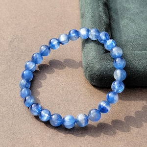 Peut inclure: Un bracelet circulaire composé de nombreuses perles sphériques polies dans des tons variés de bleu translucide. Les perles présentent une texture naturelle, fibreuse et chatoyante, reflétant la lumière sur leurs surfaces. Le bracelet est disposé en anneau.
