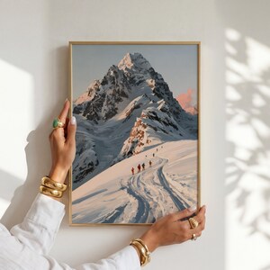 Könnte beinhalten: Ein rechteckiger Wandkunstdruck mit einem Gemälde eines schneebedeckten Berggipfels unter einem blassen Himmel. Eine Gruppe kleiner Figuren in bunter Ausrüstung überquert einen verschneiten Pfad im Vordergrund, mit tiefblauen Schatten und rosa Licht.