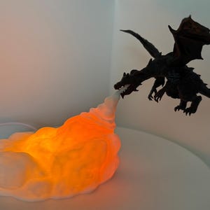 May include: A black dragon figurine with outstretched wings appears to breathe fire. The fire is a glowing orange and white cloud-like shape. The dragon is positioned above the fire, creating a fantasy scene.