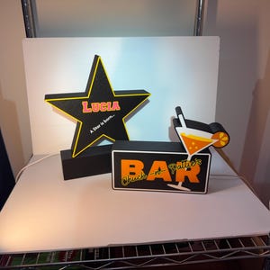 May include: A black star-shaped light with yellow trim featuring the text "LUCIA" and "A Star is born..." alongside a rectangular black light box with the text "Chuck-n-Pattie's BAR" and a graphic of an orange cocktail with a slice of fruit.