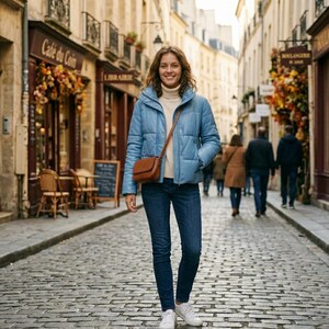 May include: A light blue puffer jacket worn over a cream-coloured turtleneck sweater, paired with dark blue denim jeans and white trainers. A small brown leather crossbody bag with a thin strap is worn across the torso on a cobblestone street.