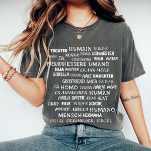 Könnte beinhalten: Dunkelgraues T-Shirt mit weißen Wörtern für 'Frau' in verschiedenen Sprachen. Die Person trägt eine goldene Halskette und hat lange, wellige Haare. Der Text enthält Wörter wie 'Tochter', 'Mutter' und 'Schwester'.
