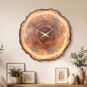 May include: A round wall clock featuring a natural wood slice design with visible tree rings in shades of brown and tan. The clock face includes white numerals at the 12, 3, 6, and 9 positions, with thin gold-colored hour and minute hands.