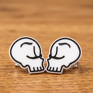May include: A pair of stud earrings featuring white skull shapes with black outlines and stylised closed eyes. The skulls are oriented to face each other, showcasing a smooth finish and a simple, graphic design with clear black line details.