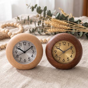 May include: Two round analog clocks with wooden frames. The left clock has a light wood frame and a silver face with black numerals. The right clock has a dark wood frame and a gold face with black numerals. Both feature black hands.