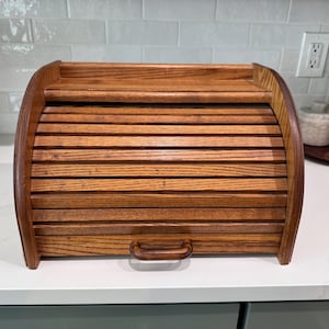 May include: A medium-toned wooden bread box featuring a roll-top tambour door with horizontal slats and a small, curved handle. The structure has a flat top surface and arched side panels, providing a secure enclosure for bread storage.