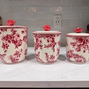 May include: A set of three cream-colored ceramic canisters featuring a red floral pattern. Each canister has a matching lid topped with a sculpted red rose handle. The canisters vary in size, with the largest on the left and smallest on the right.