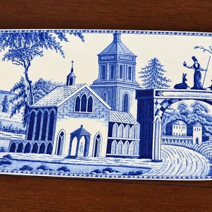 May include: An elongated oval ceramic serving platter featuring a blue and white transferware pattern. The design depicts a landscape with a church, a stone archway, a sailboat on water, and trees. The border has a repeating geometric motif.