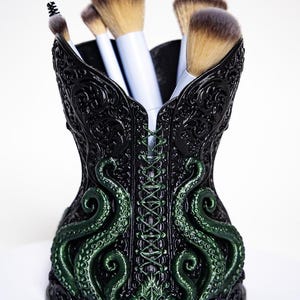 Peut inclure: Un porte-pinceaux de maquillage en forme de corset noir avec des motifs en relief complexes et des tentacules verts en relief sur le devant. Le support contient plusieurs pinceaux de maquillage à manches blancs et poils bruns.