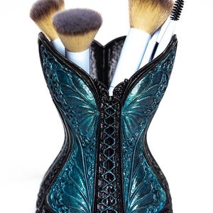 Peut inclure: Un porte-pinceaux de maquillage en forme de corset noir et bleu sarcelle, orné de motifs d'ailes de papillon en relief et d'un laçage central. Le récipient contient quatre pinceaux de maquillage à manches blancs et poils bruns.