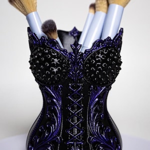 Peut inclure: Un porte-pinceaux de maquillage en forme de corset noir et violet avec des motifs en relief complexes, un laçage central et deux bonnets texturés à picots. Plusieurs pinceaux à maquillage aux manches clairs sont rangés à l'intérieur.