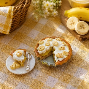 Pode incluir: Uma torta de creme e banana em miniatura com uma fatia cortada, coberta com chantilly e fatias de banana. Um prato pequeno com uma fatia de torta e uma colher estão ao lado da torta. Uma cesta de bananas e uma tigela de chantilly estão no fundo.