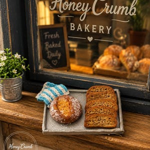 May include: Miniature bakery scene with a window display. A small chalkboard sign reads "Fresh Baked Daily." A tray holds a round loaf, sliced bread, and a blue and white striped cloth. The bakery is named "Honey Crumb."
