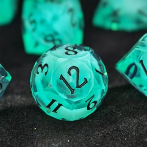May include: A translucent, bright teal twelve-sided die featuring sharp, faceted edges and bold, black engraved numbers. The die displays the number twelve prominently in the center, surrounded by other visible digits including eight, three, six, and eleven.
