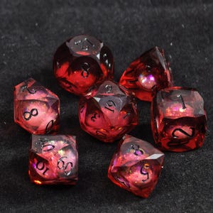 May include: A set of seven polyhedral dice made of translucent red resin with black ink numbers. Each die contains embedded iridescent pink and gold glitter particles that catch the light, creating a shimmering effect within the faceted shapes.