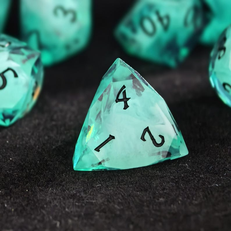 May include: A four-sided translucent teal polyhedral die with sharp edges and black engraved numbers. The die features a cloudy, swirling internal pattern and is positioned among other similar dice in the background, showcasing its geometric shape.