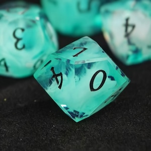 May include: A translucent teal and white swirled polyhedral ten-sided die featuring sharp edges and bold black engraved numbers. The die displays a cloudy, smoke-like internal pattern with a glossy finish, showing the numbers seven, four, and zero.