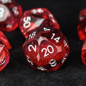 May include: A twenty-sided die made of translucent red resin with embedded glitter particles. The die features white engraved numbers, with the number 20 prominently displayed in the center. Other visible numbers include 2, 8, 14, and 18.