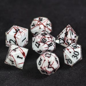 Peut inclure: Un ensemble de sept dés polyédriques présentant une base blanche avec un motif d'éclaboussures rouges et des chiffres gravés en noir. La collection comprend un d20, d12, d10, d8, d6, d4 et un dé de pourcentage, avec une finition extérieure claire.