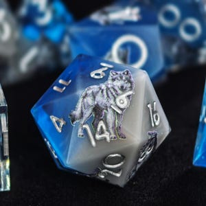 May include: A twenty-sided die featuring a gradient of deep blue to opaque gray resin. The surface displays a detailed illustration of a standing wolf in shades of gray and white. The numbers are engraved in a metallic silver finish, clearly visible.